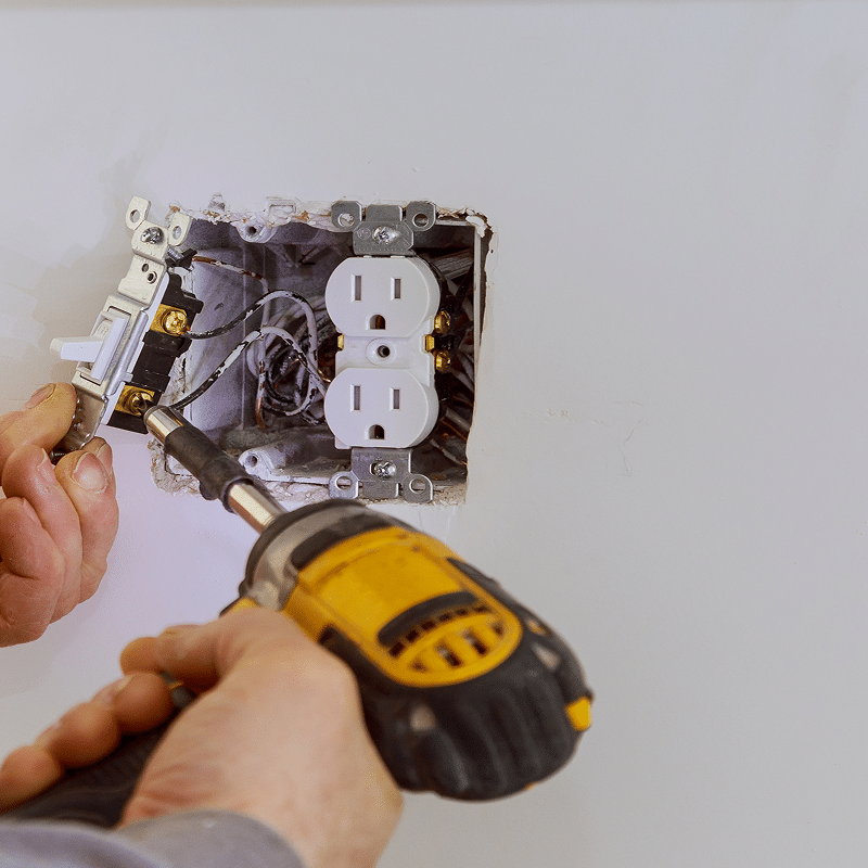 Mains d'électricien installant une prise électrique double et un interrupteur mural avec un tournevis électrique, câblage visible.