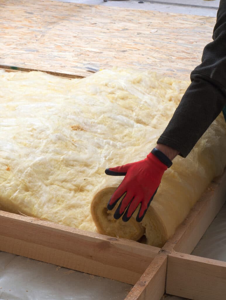 Main gantée rouge déroulant une couverture de laine minérale jaune pour isoler un plancher en bois avec des chevrons.