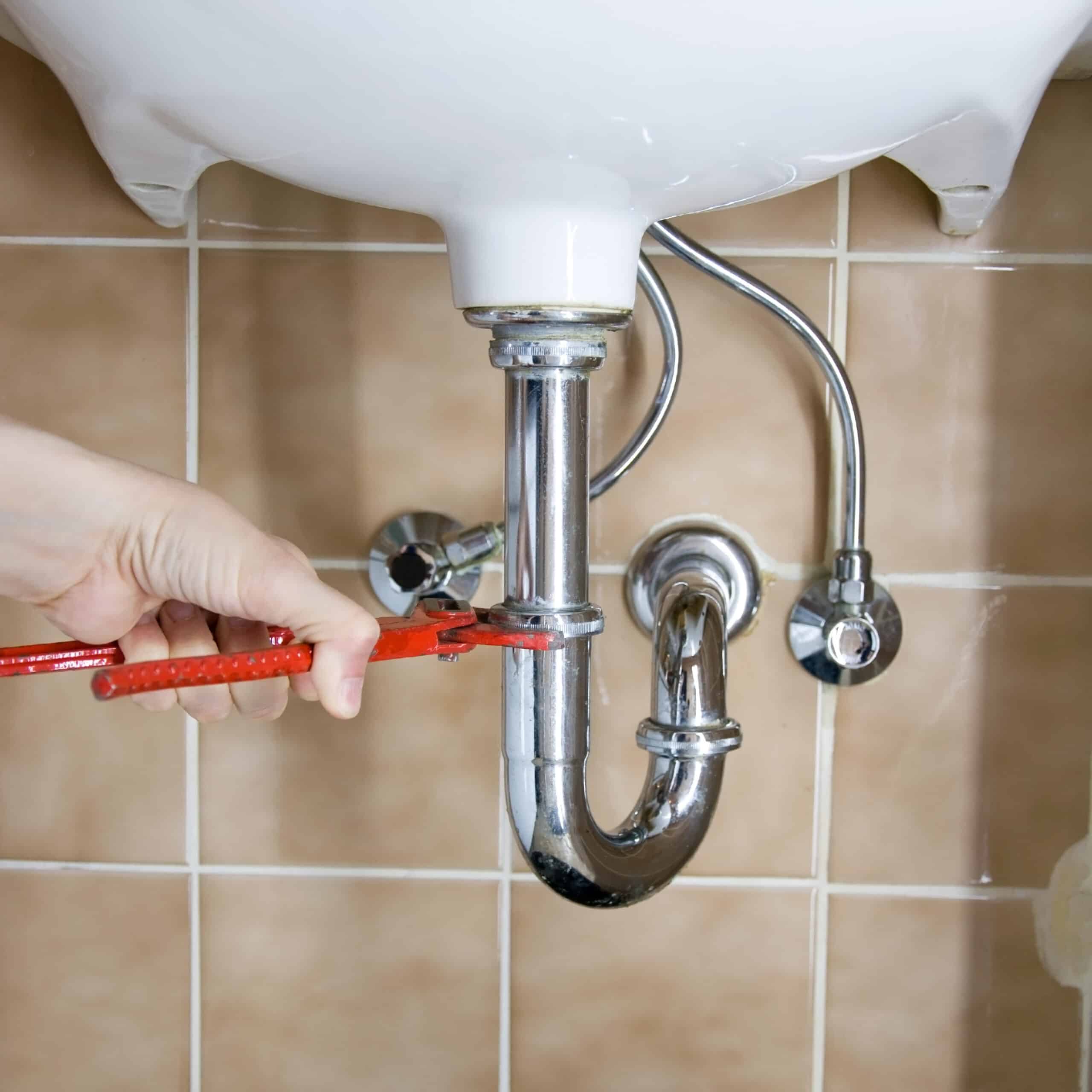 Main serrant un siphon d'évier chromé avec une clé à molette rouge. Mur carrelé beige en arrière-plan.