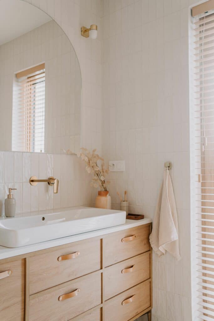 Salle de bain moderne: vasque blanche, meuble en bois clair, robinet mural doré, miroir arqué. Mur carrelé blanc, stores clairs, détails naturels.